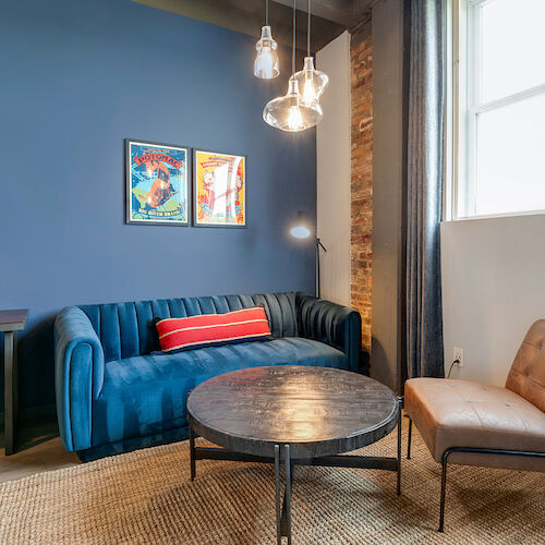 A modern living room with a blue couch, brown chair, round coffee table, and two wall art pieces, illuminated by three hanging lights.