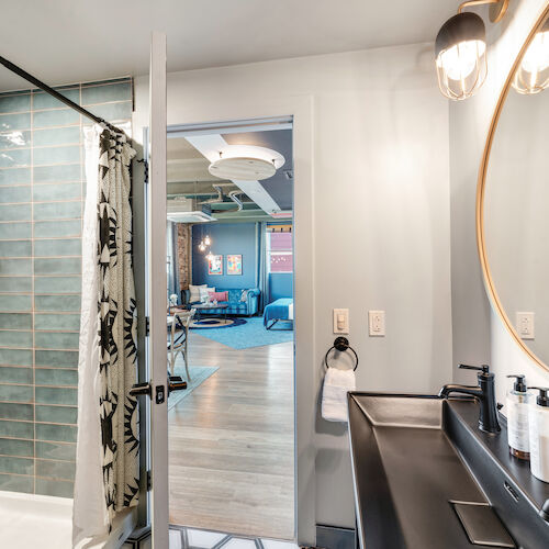 The image shows a bathroom with a shower area, blue tiles, and a vanity with a round mirror. The bathroom opens into a living area with modern furniture.