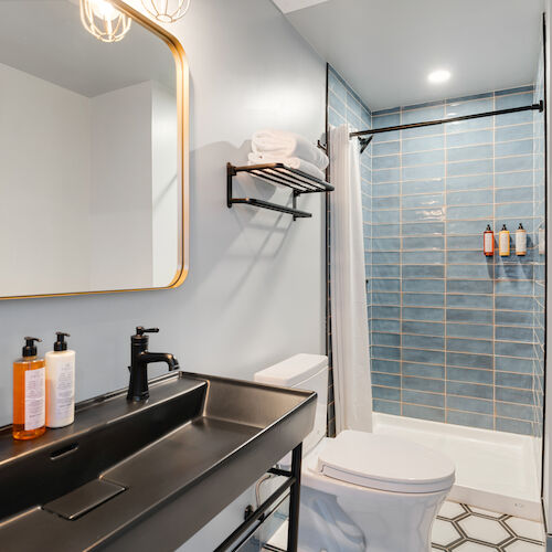 A modern bathroom with a large mirror, dual sink, white countertop, toilet, and a shower with blue subway tiles.