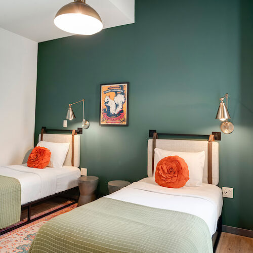 The image shows a bedroom with two twin beds, green walls, orange accent pillows, wall lamps, a decorative picture, and a large window covered by curtains.