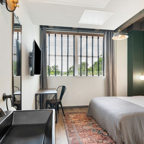 A modern hotel room with a bed, TV, desk, chair, and a window. The decor includes a rug, a wall mirror, and a sink. The room appears well-lit.