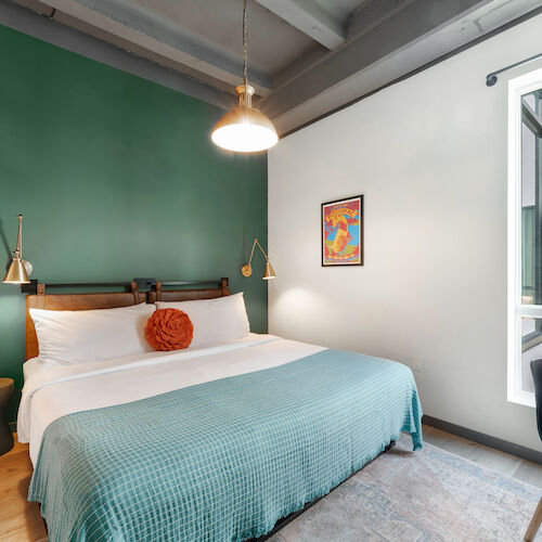 A cozy bedroom with a large bed, green accent wall, modern lighting, framed art, chair, desk, and a window providing natural light ends the sentence.