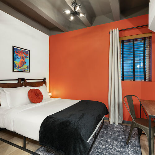 A cozy bedroom with an orange accent wall, bed with white and black linens, artwork, window, desk, chair, and wall-mounted TV.