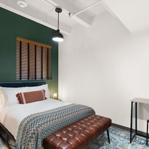 A modern bedroom with a single bed, green accent wall, leather bench, side tables, chair, desk, and framed artwork on a white wall.