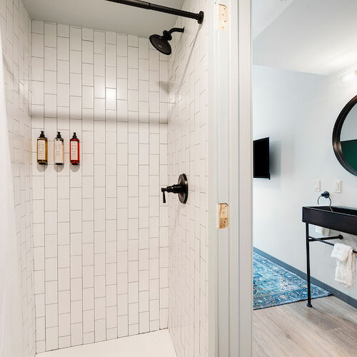 A modern bathroom with a tiled shower, three mounted bottles, a black faucet, a black vanity, a round mirror, and a wall-mounted TV.