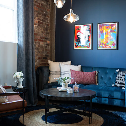 A cozy living room with a blue tufted sofa, colorful wall art, a round coffee table, hanging lights, and a mix of decor pieces.