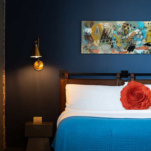 The image shows a bedroom with a bed, blue walls, abstract art, a red throw pillow, and a wall-mounted light fixture.