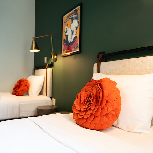 The image shows a bedroom with two single beds, orange flower cushions, white bedding, a green wall, and a wall-mounted lamp.