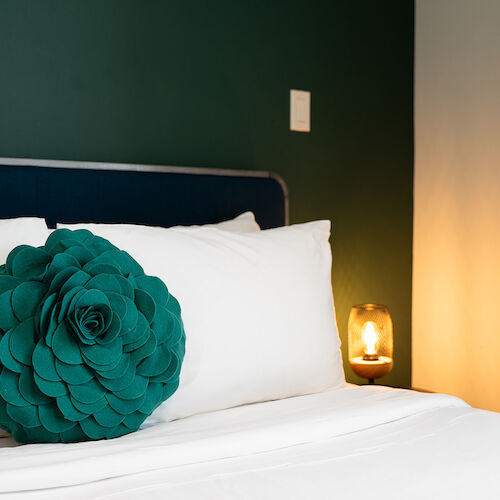 A neatly-made bed with white linens and a decorative green flower-shaped pillow, next to a lit bedside lamp, is shown in the image.