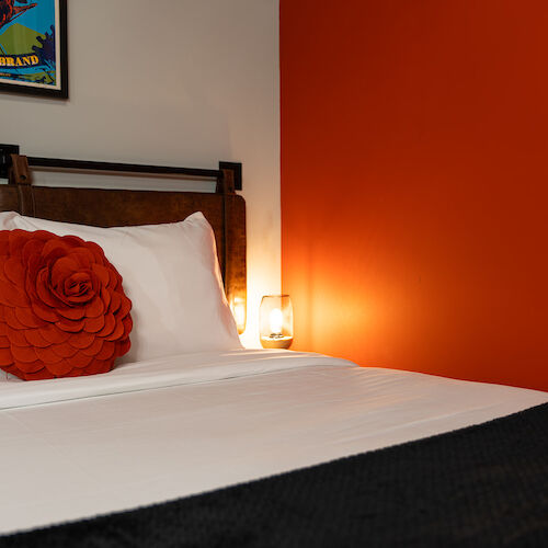 A neatly made bed with white sheets and a red rose-shaped cushion, beside a lamp on a bedside table and an orange wall in the background.