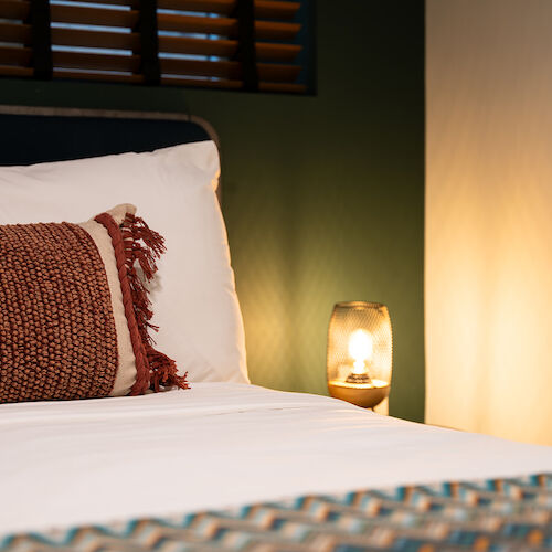 A cozy bedroom scene with a bed, a decorative pillow, and a soft-lit lamp on a nightstand against a dark green wall and window shutters.