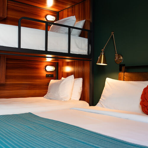 A cozy room with a bunk bed and a single bed, white linens, a red cushion, wooden accents, and a wall-mounted reading light.