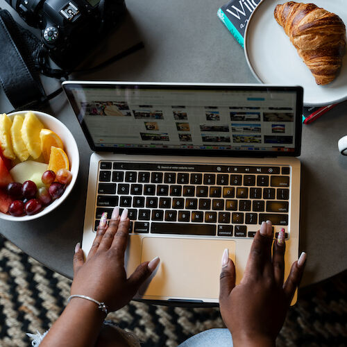 A person is using a laptop on a table with a bowl of fruit, a croissant, a cup of coffee, and a camera placed nearby.