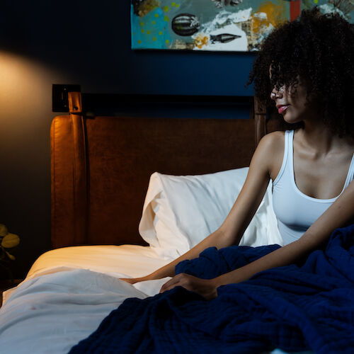 A person with curly hair sits in a bed covered with a blue blanket in a dimly lit room featuring wall art and a small side table with a few items.