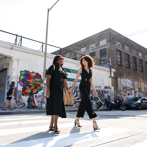 Two people are walking across a street in an urban area with graffiti art on buildings in the background. The sky is clear and sunny.