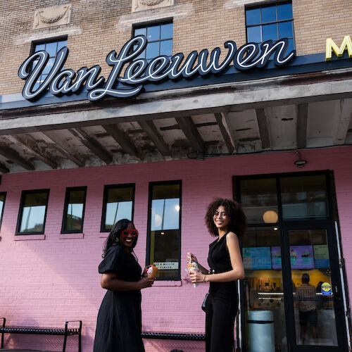 Two women stand outside Van Leeuwen ice cream shop, smiling and holding ice creams, against a building with a pink exterior and a vintage sign.