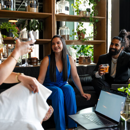 Three people are socializing in a cozy setting, with drinks and a laptop on the table, surrounded by plants and shelves with various items.