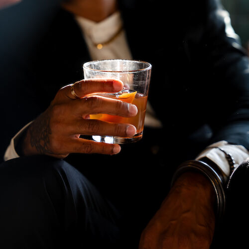 A person holding a glass of an amber-colored drink with ice and an orange slice, seated and dressed in dark clothing.