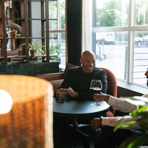 Two people are sitting at a table in a cozy indoor setting with drinks, smiling and conversing by the window.