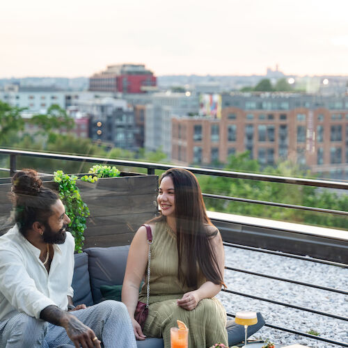 A man and woman are sitting on an outdoor patio with food and drinks on a table, enjoying a view of city buildings.