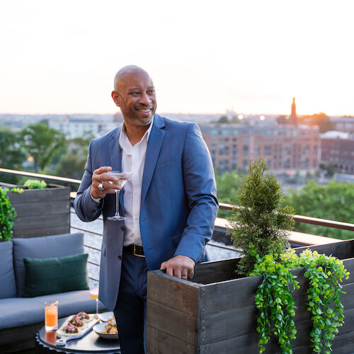 A man in a blue suit is holding a drink on a rooftop patio, with greenery and cityscape in the background during sunset.