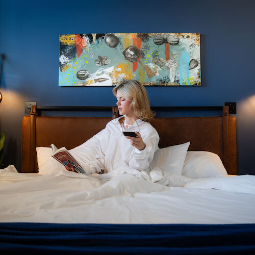 A person is sitting in bed with a book and a drink, surrounded by modern decor and plants, under a colorful abstract painting on a navy-blue wall.