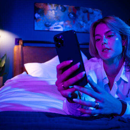 A person is lying on a bed in a dimly lit room, looking at their phone, with blue and pink lighting illuminating the scene.