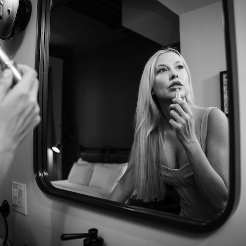 A woman is looking at a mirror while applying lipstick with one hand, captured in black and white. She appears to be in a bedroom or dressing area.