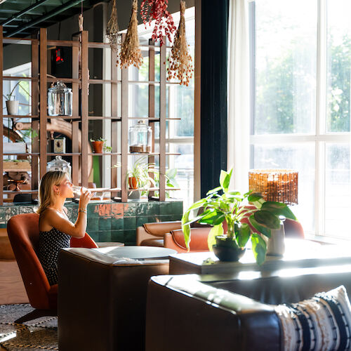 A lounge area with cozy seating, a woman sipping a drink by the window, warm lighting, plants, and decorative shelving in the background.