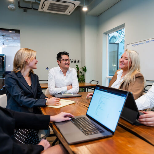 A group of five people are sitting around a table in a meeting room, engaged in discussion with laptops and notebooks in front of them.