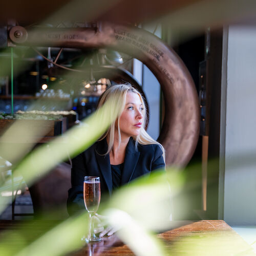 A woman sits at a table with a drink in hand, surrounded by green plants and modern decor in a café or restaurant setting.