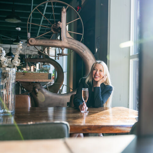 A person sits at a wooden table in a modern, rustic space with large windows, holding a glass of champagne and smiling, with decor in the background.