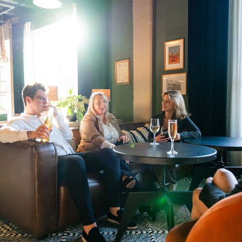 Four people are sitting in a cozy, well-lit lounge area, talking and enjoying drinks around a round table.