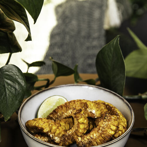 A white bowl of seasoned corn on the cob with a lime wedge, surrounded by green leaves, is placed on a dark surface.