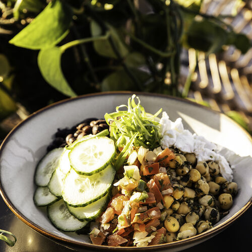 A bowl with sliced cucumbers, chopped tomatoes, chickpeas, beans, and shredded coconut, garnished with green herbs, sitting on a dark surface.