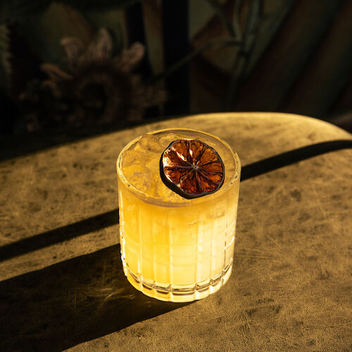 A yellow drink in a glass, garnished with a dried citrus slice, sits on a table in warm lighting.