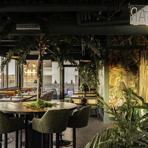 A stylish indoor restaurant with green plants, modern furniture, and large windows for natural light, creating a cozy and inviting atmosphere.