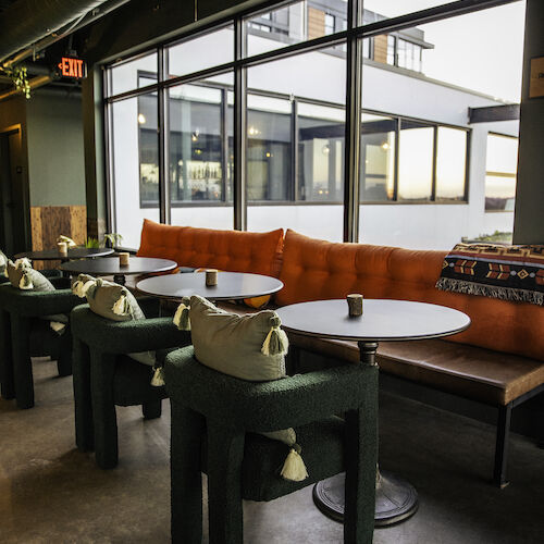 The image shows a modern cafe with round tables, chairs, and an orange cushioned bench, located near large windows letting in natural light.