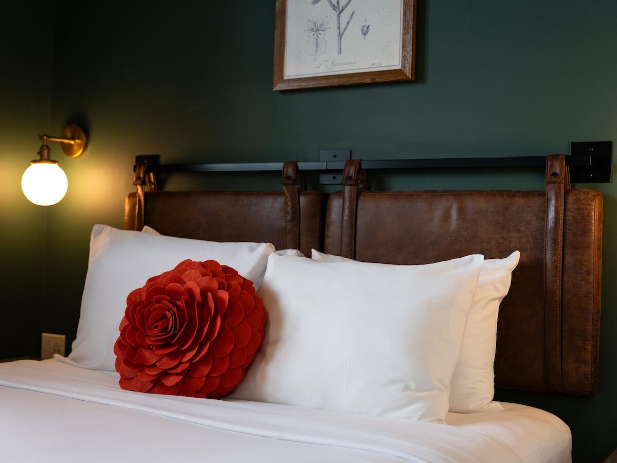 A cozy bed with white pillows, a red flower cushion, a brown headboard, green walls, two wall-mounted lamps, and a framed artwork above.
