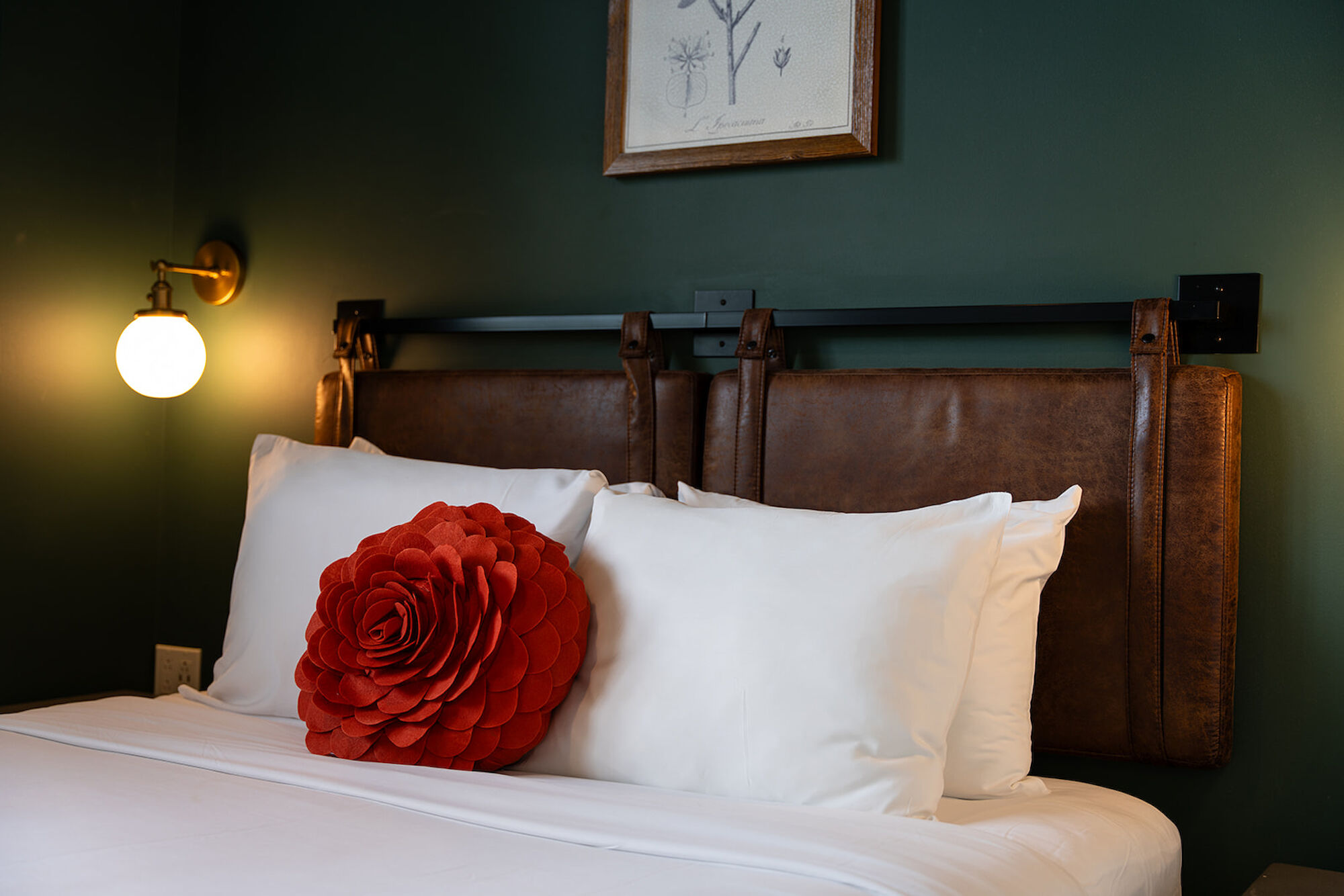 A cozy bedroom setup featuring a bed with white linens, a large red flower-shaped pillow, a leather headboard, and wall-mounted lights.