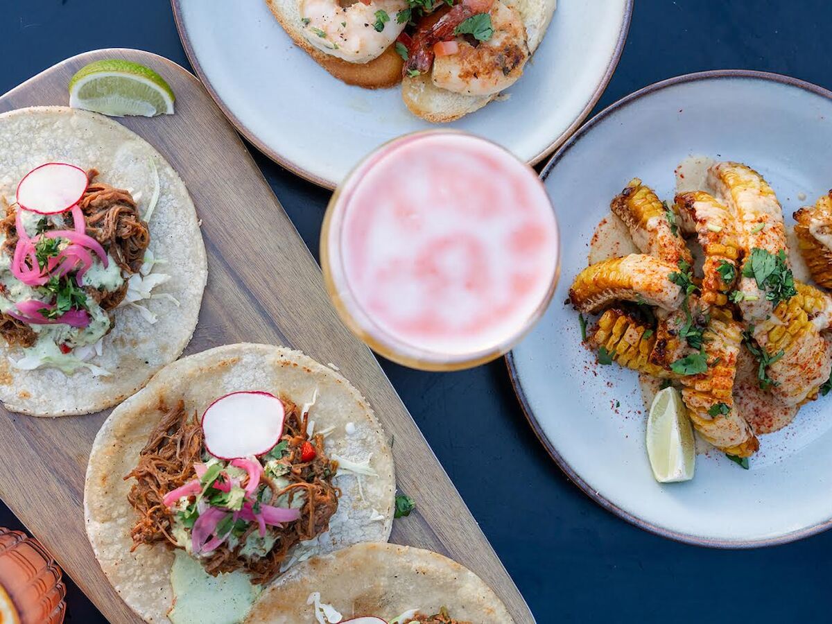 A spread of tacos on one plate, grilled corn on another, and a pink drink in the center, garnished with lime on the side.