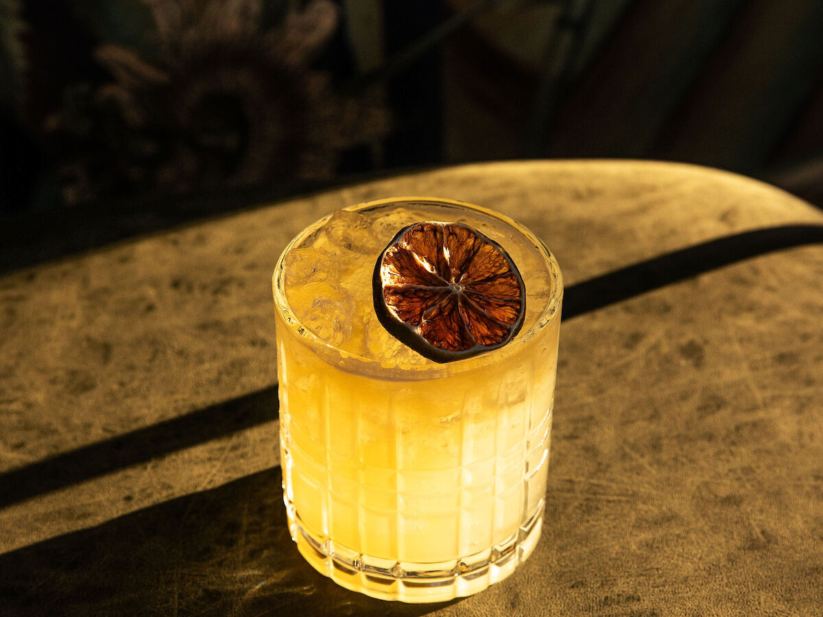 A glass of yellow cocktail garnished with a dried citrus slice, set on a surface with light casting shadows across it.