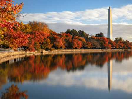 A scenic view of a lake with autumn-colored trees and a prominent obelisk monument reflecting in the calm water under a partly cloudy sky.