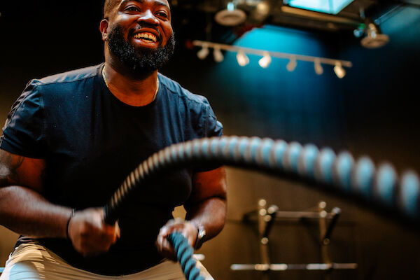 A person is exercising with battle ropes in a gym, smiling and wearing a black shirt and shorts in a well-lit space.