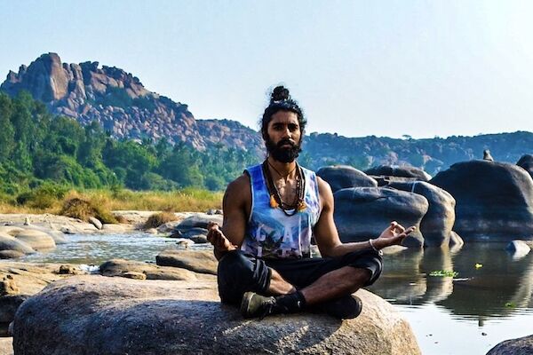 A person is meditating on a large rock near a river, with hills and greenery in the background.