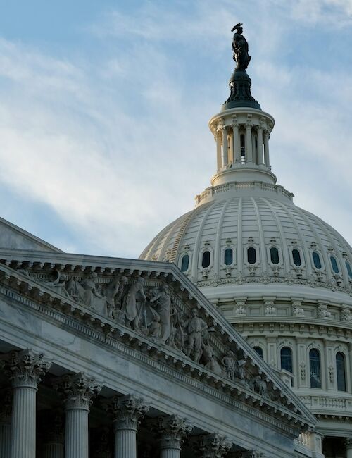 The image shows the dome of the U.S. Capitol building, featuring classical architecture with columns and detailed carvings, under a blue sky.