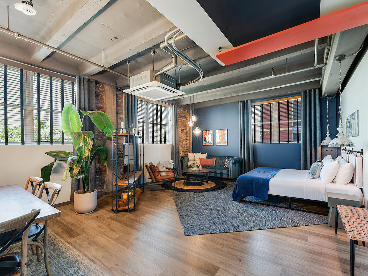 A modern loft with a cozy bedroom, living area, plants, and stylish decor, featuring large windows and industrial design elements.