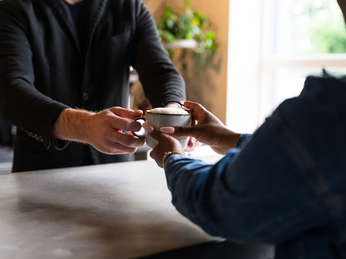 A person is handing a cup with foam on top to another person across a countertop in a cozy setting.