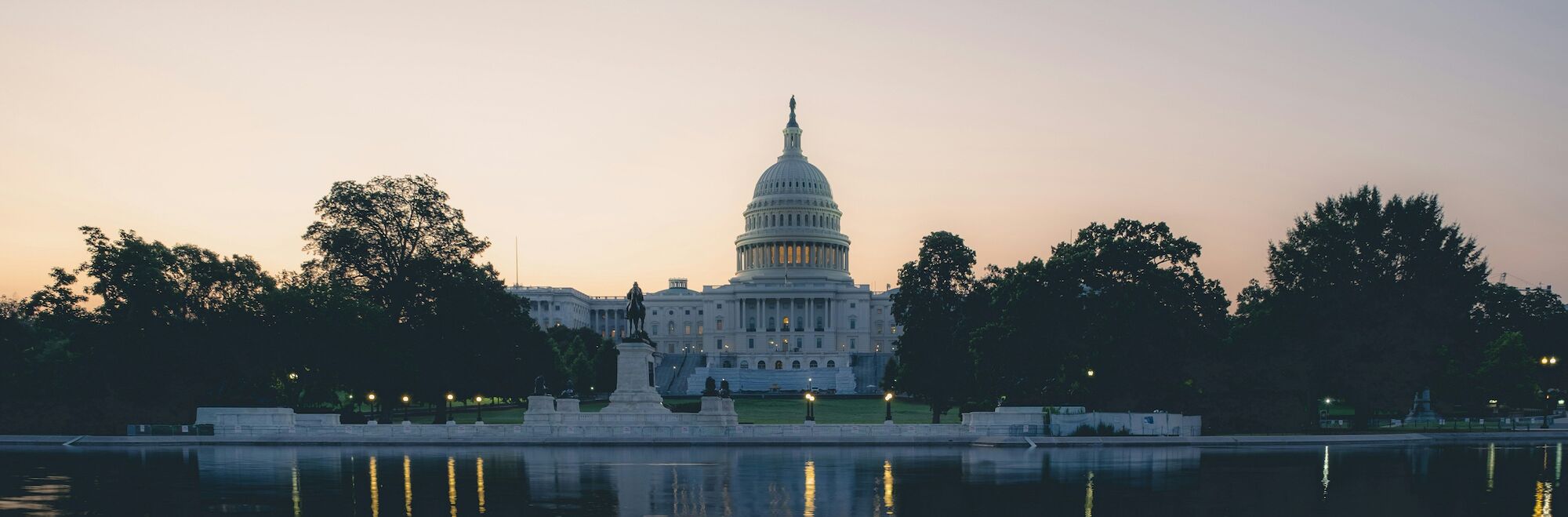 The image shows the United States Capitol building at dusk, with its reflection in a body of water in the foreground.