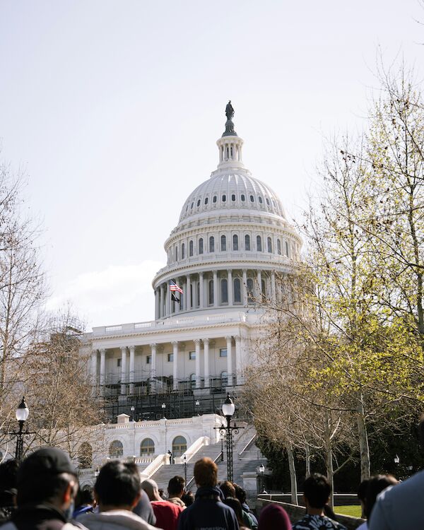 The image shows the U.S. Capitol building with people in the foreground, surrounded by leafless trees under a clear sky.